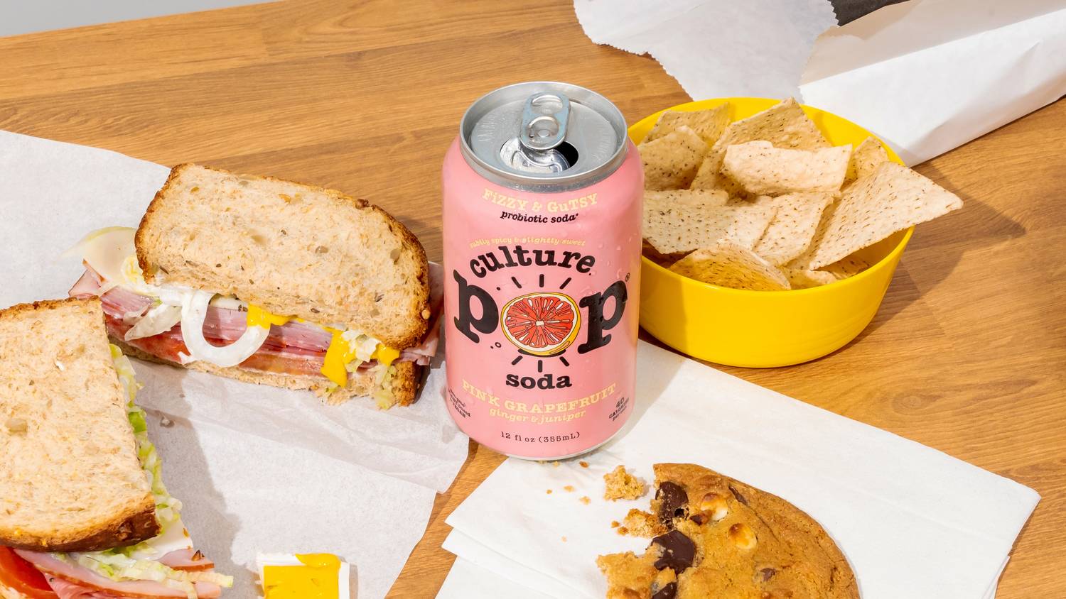Deli sandwich spread with a can of Culture Pop soda, tortilla chips, and a chocolate chip cookie