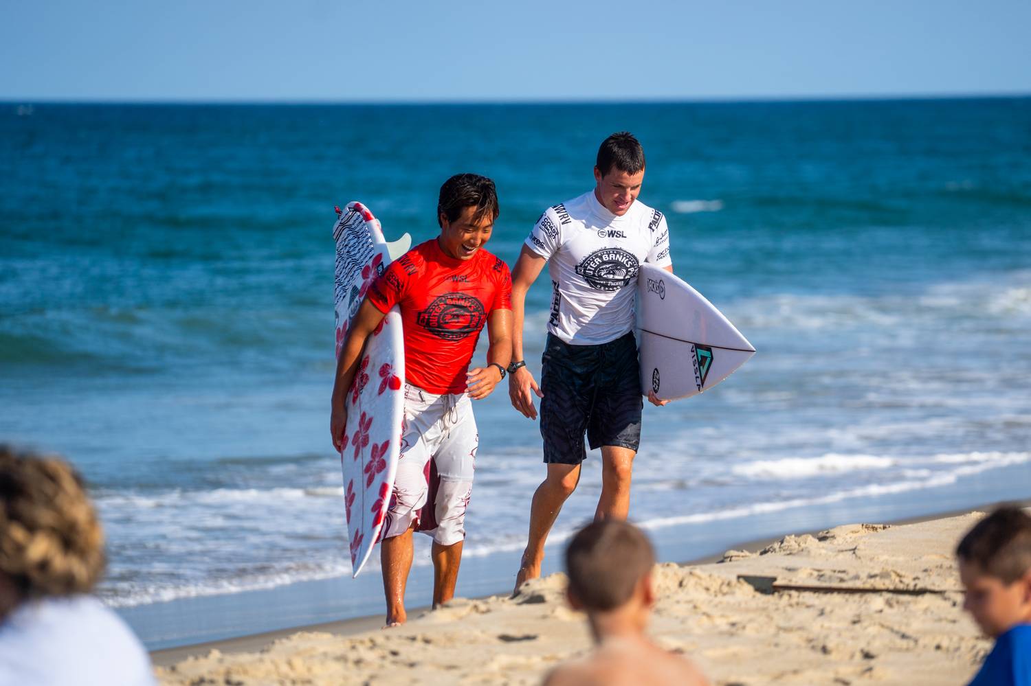 Two competitive surfers carrying boards and laughing on the beach