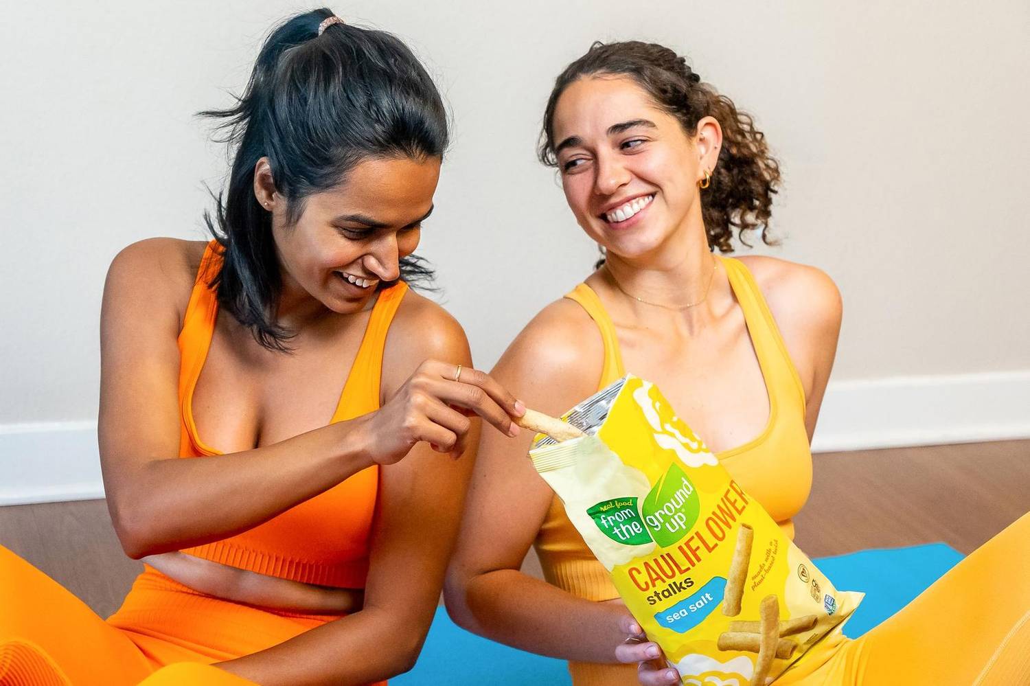 Two women in workout gear sharing a bag of cauliflower snack sticks