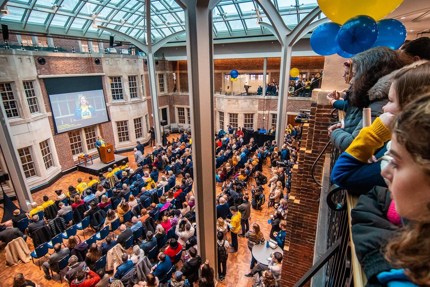 Crowd gathered in a balloon-decorated atrium watching a speaker on screen