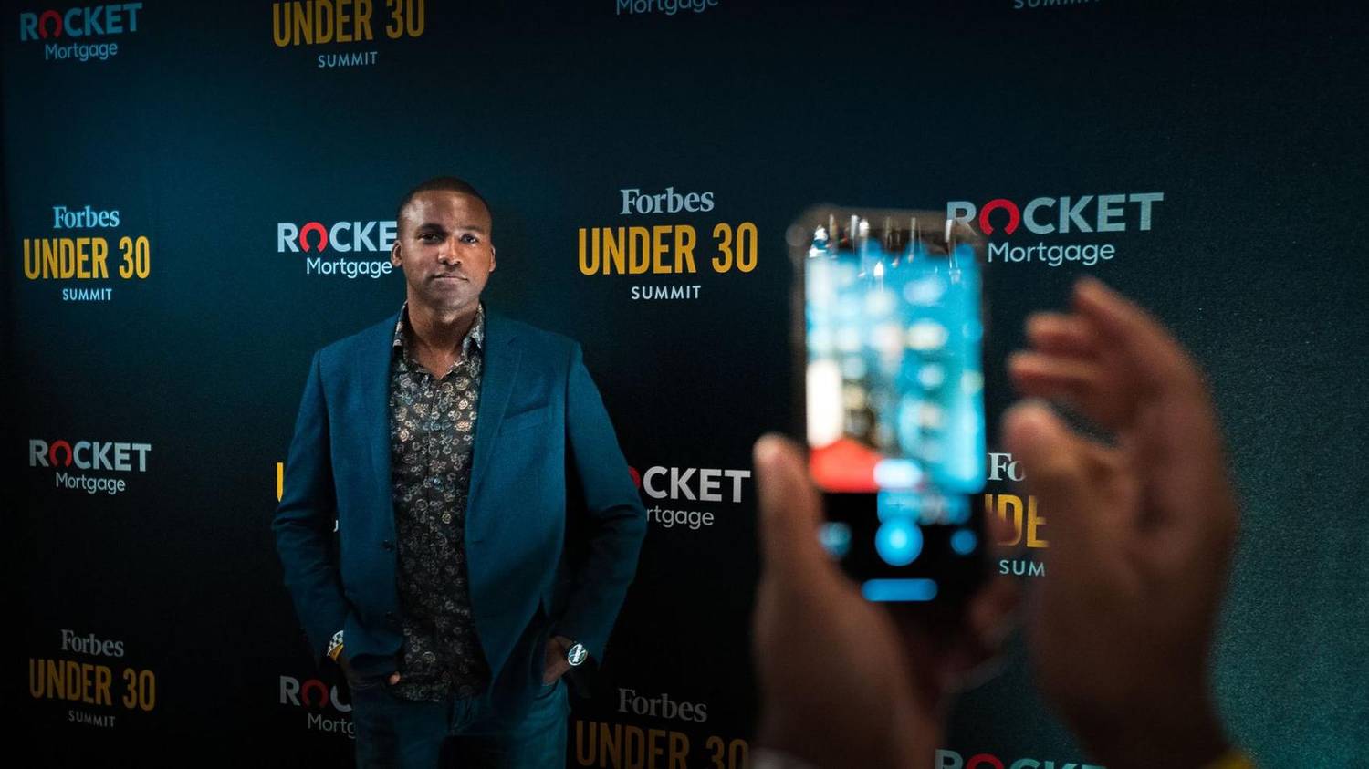 Man posing for a photo at a Forbes Under 30 Summit step-and-repeat backdrop