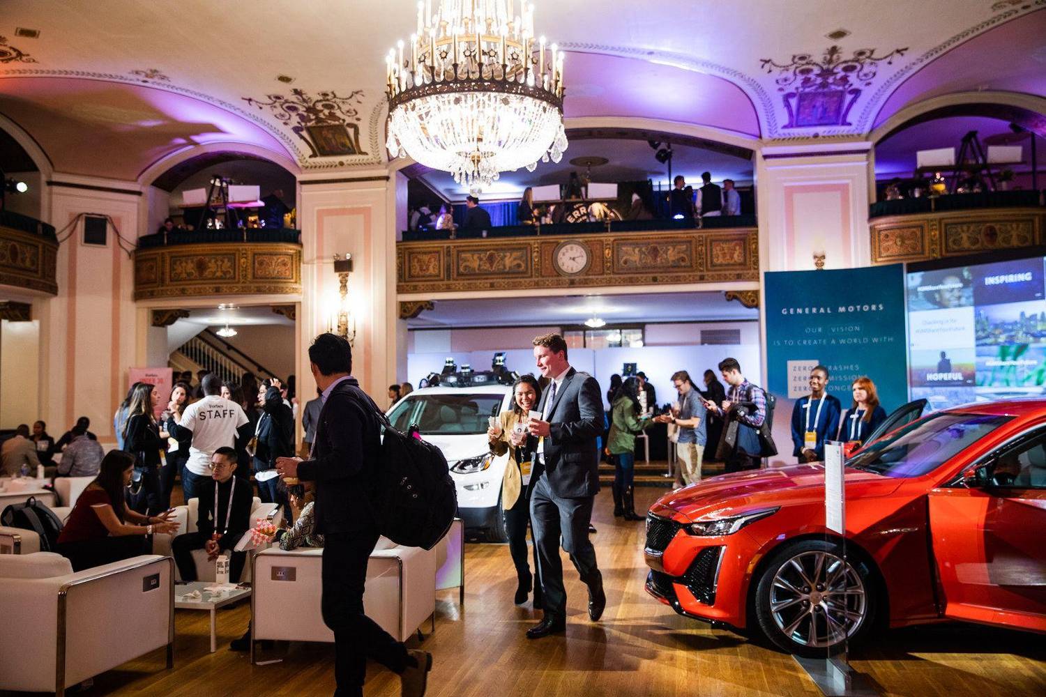 Guests mingling in a grand ballroom lounge beside vehicles on display at an auto show gala