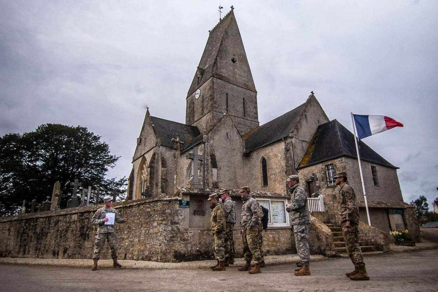 Soldiers gathered outside a stone church in Normandy beneath a French flag