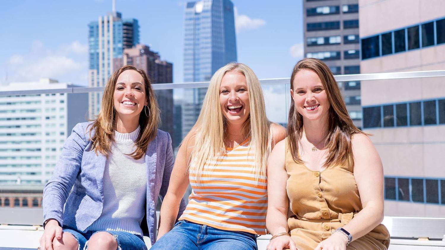 Three women laughing together on a rooftop with a city skyline behind them