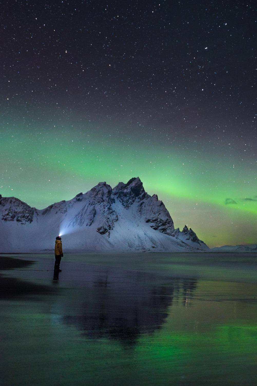 Aurora borealis over a snow-capped mountain with a figure on the shore