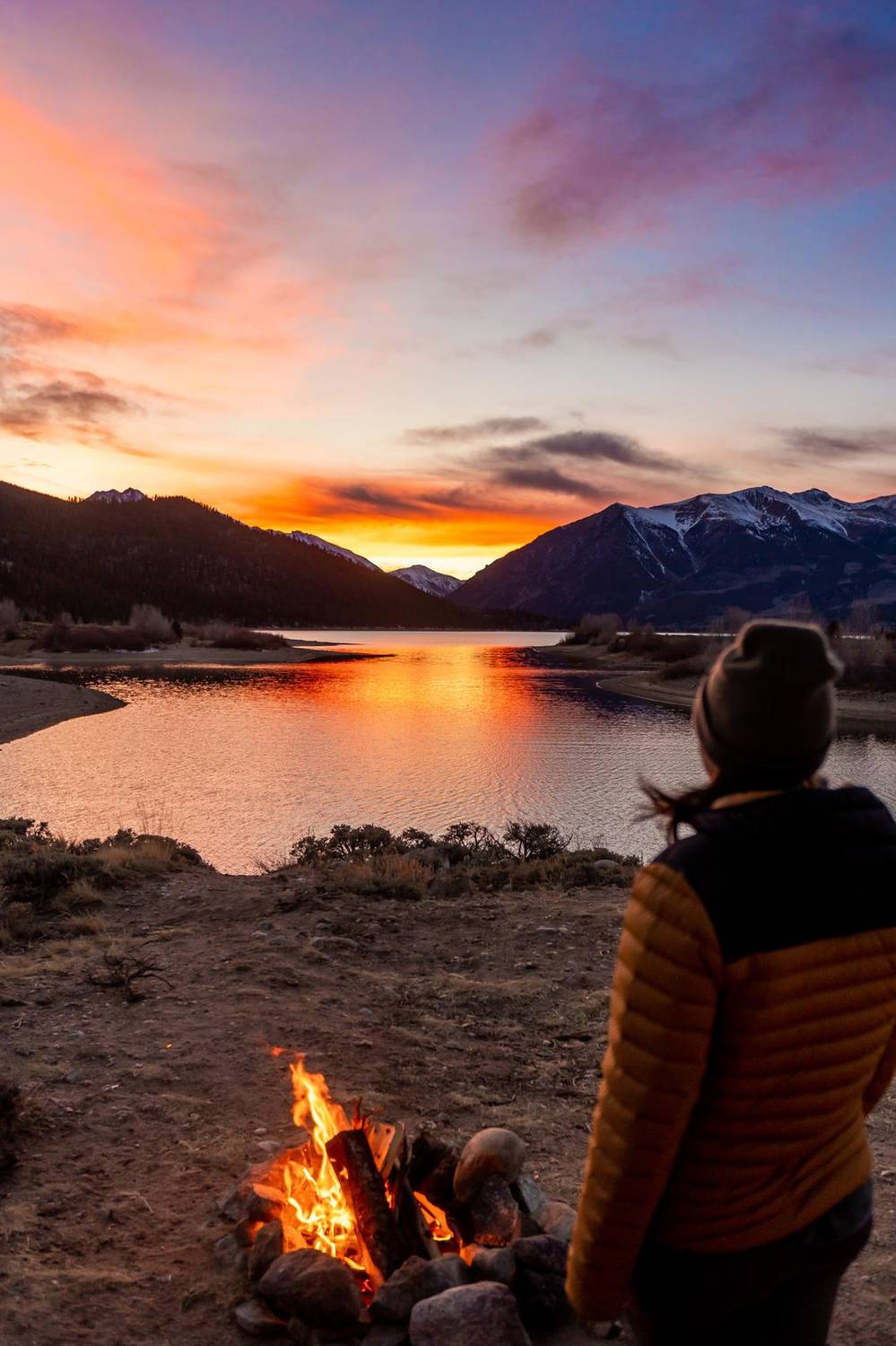 Person in a beanie warming by a campfire beside a mountain lake at sunset