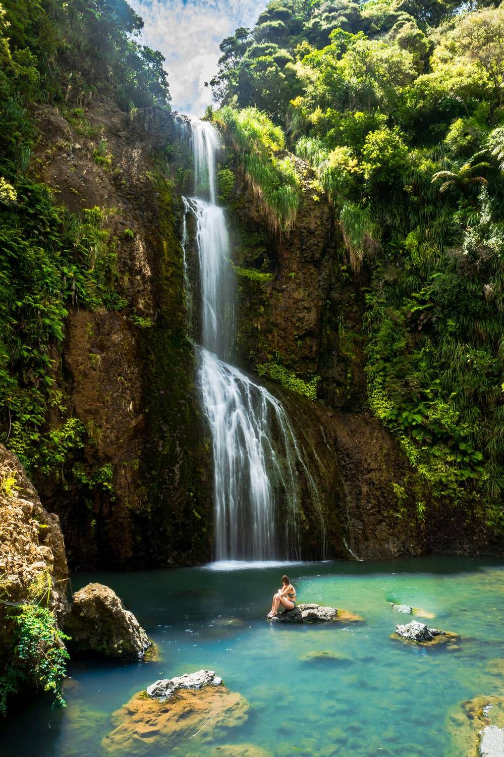 Woman sitting by a waterfall pool in a lush gorge